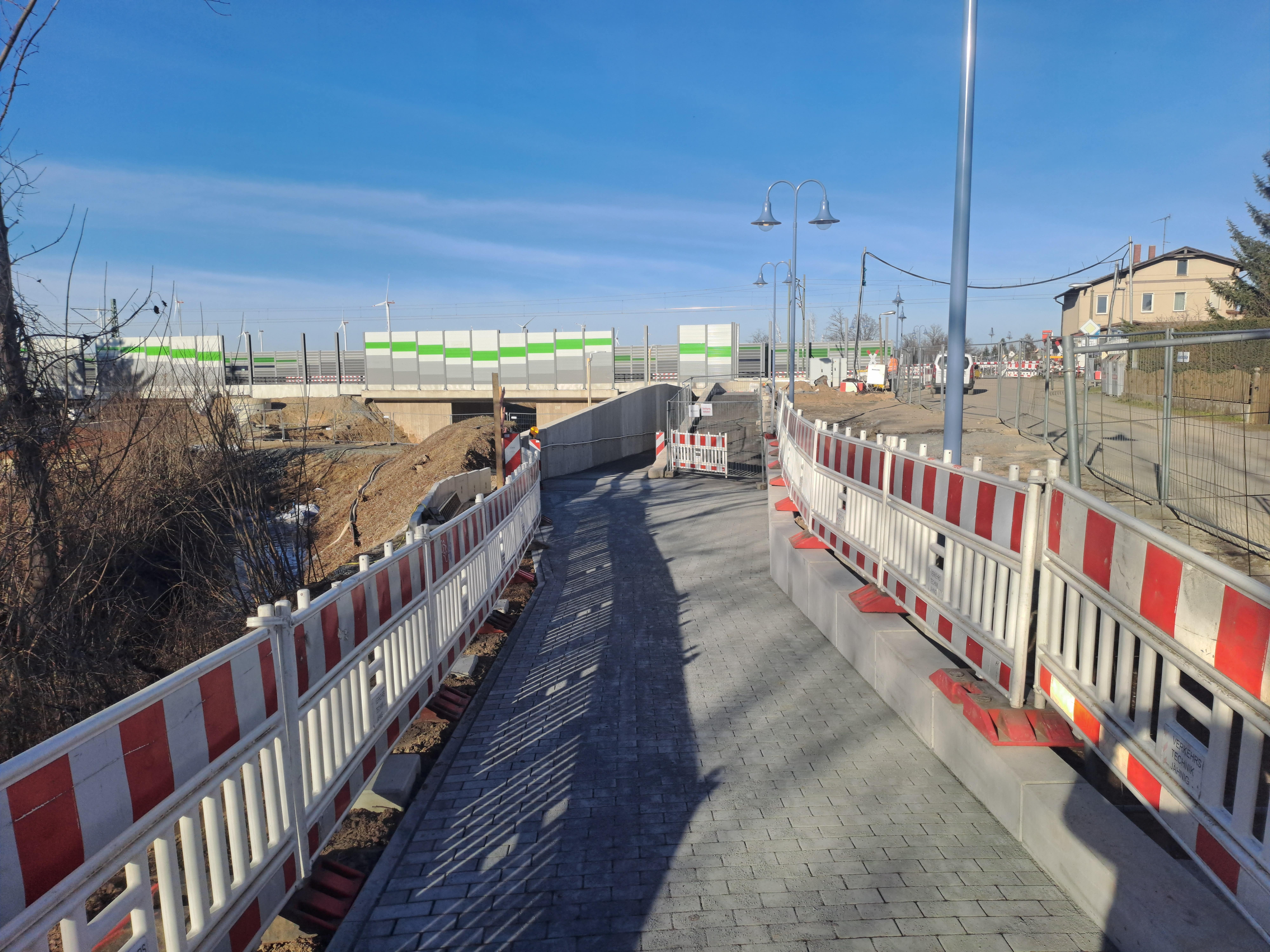 Gepflasterter Fußweg, rechts und links Bauabsperrungen, im Hintergrund Brücke mit Schallschutzwänden