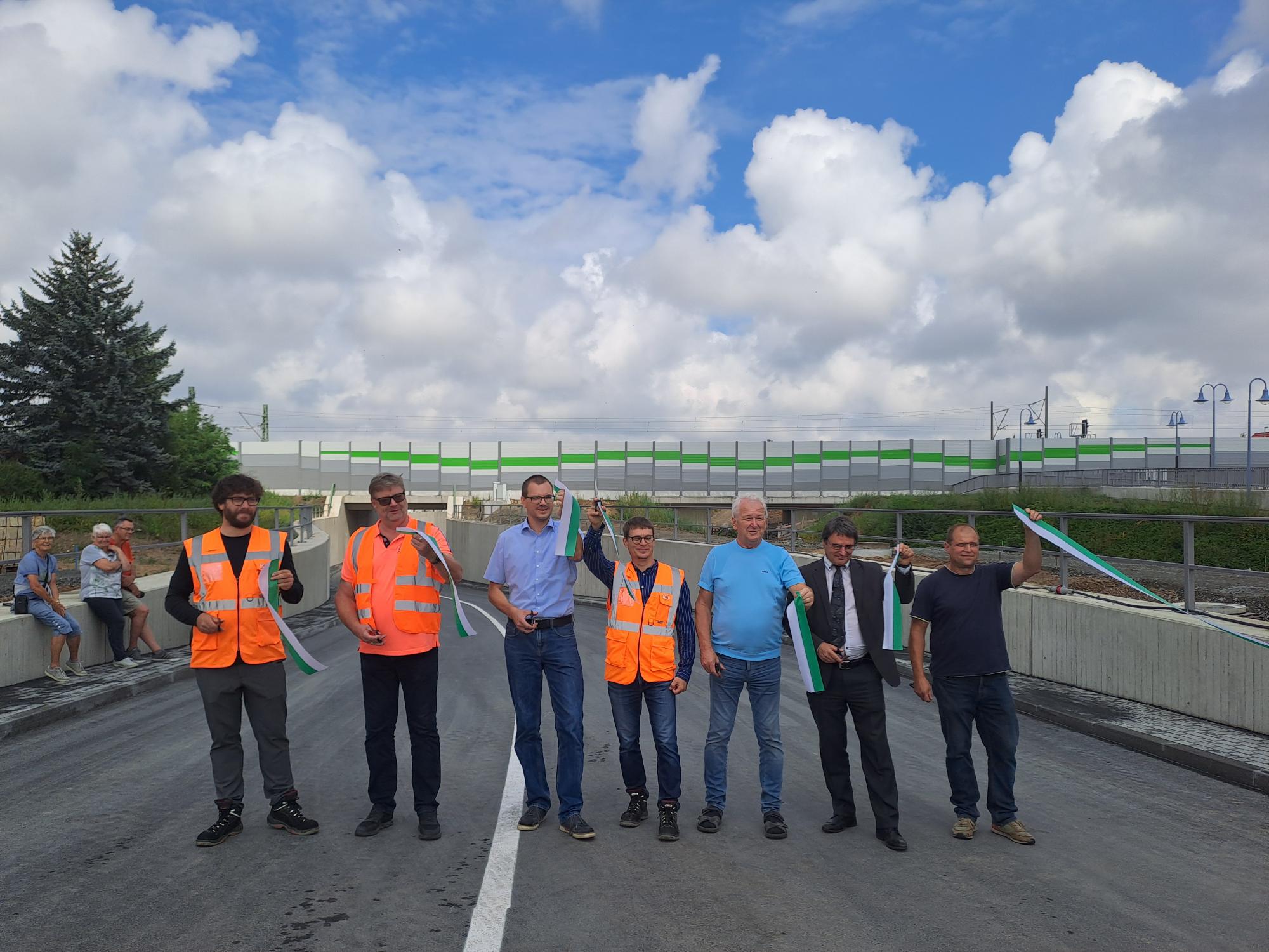 Sieben Personen stehen auf einer neu gebauten Straße und schneiden ein grünes Band durch, um die Eröffnung der Brücke zu feiern. Im Hintergrund sind Zuschauer und eine Brücke zu sehen.